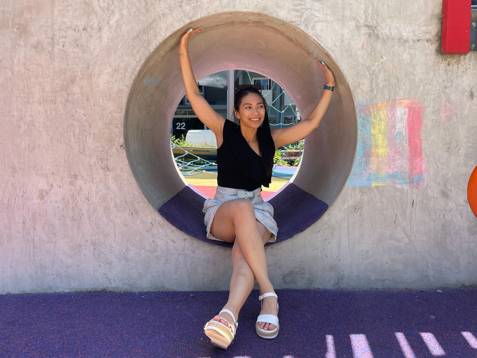 A woman wearing a black top with one shoulder, a floral white skirt and white platform sandals. She is sitting in a small tunnel that is part of a children’s playground. She has her hands reaching up and touching the roof of the tunnel.