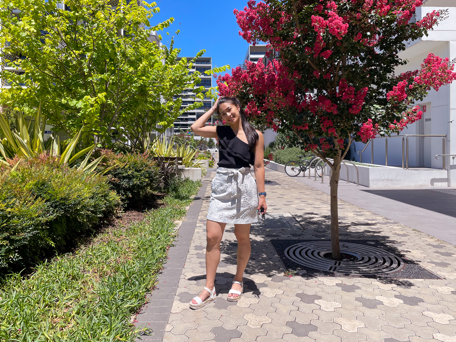 The same woman from previous photos on this page, in the same outfit. She is holding her sunglasses in her hand, and standing next to some trees with pink flowers. In the background are high-rise apartments.