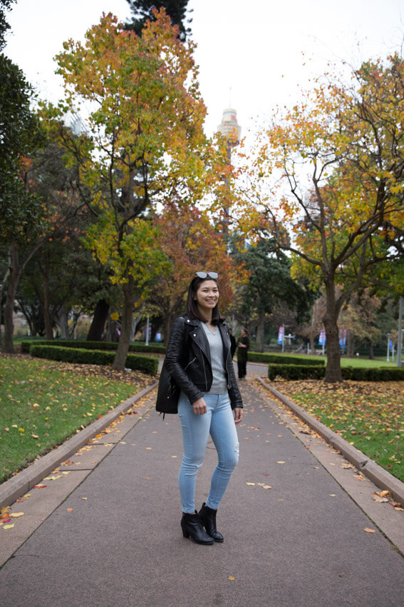 A woman with short dark hair, dressed in light blue jeans and a black leather jacket, standing on a path surrounded by trees with green-yellow leaves
