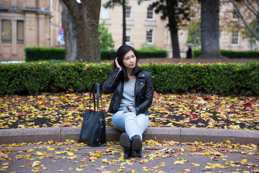 The same woman in the previous photos on this webpage, sitting on the edge of a park path with her hand held to her hair. There is a black handbag on the concrete next to her.