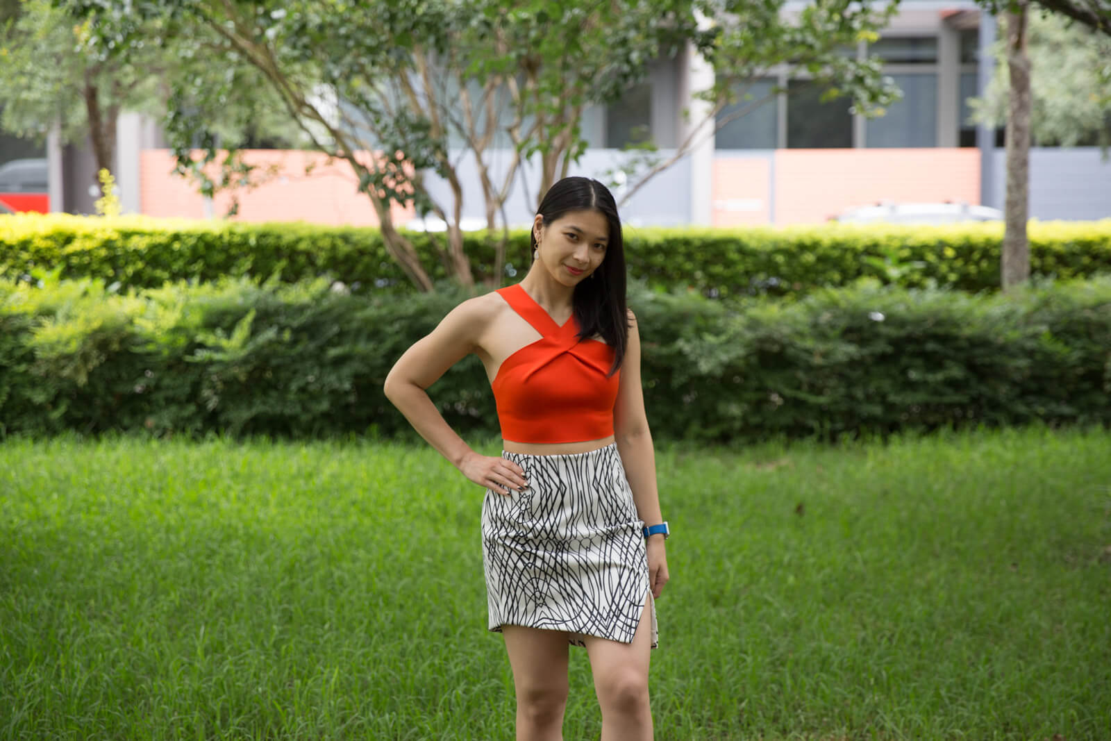 A woman with short dark hair wearing an orange crop that crosses at the neck. Her skirt is white with an abstract black print. She is standing in a very green garden with hedging in the background and has a hand on her hip.