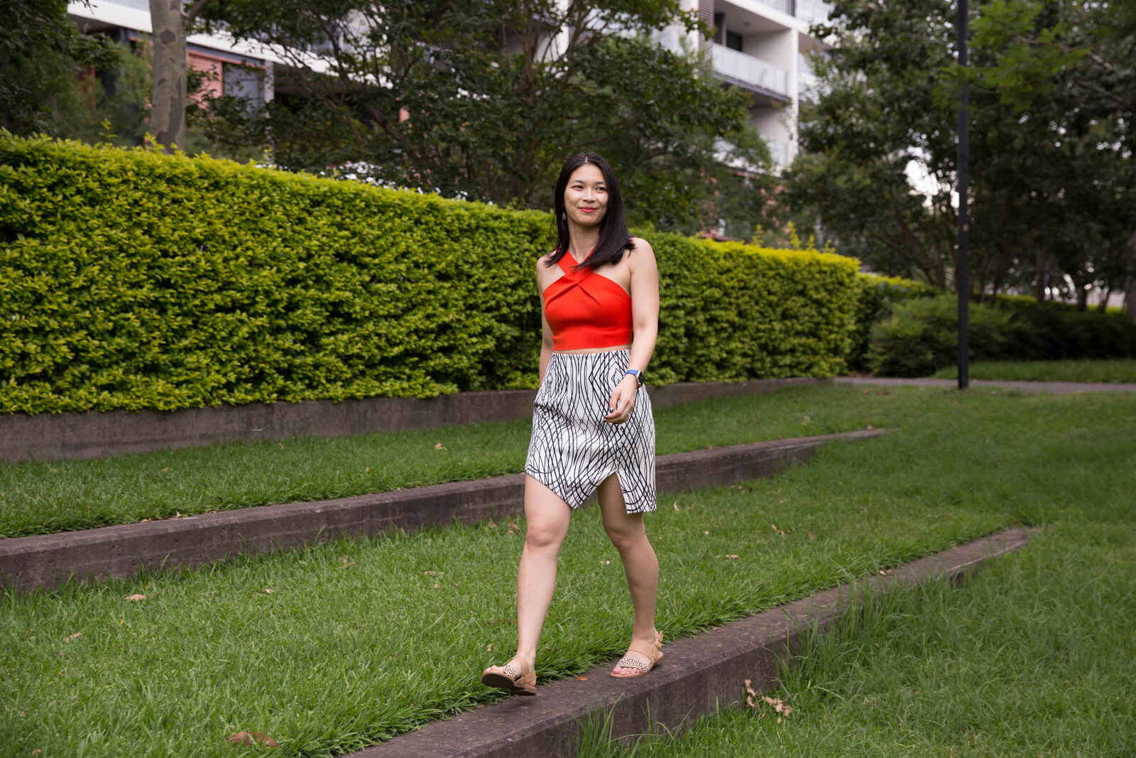 A full body shot of a woman with short dark hair, wearing an orange crop top, and black and white abstract printed skirt, walking across a concrete step lined with grass.