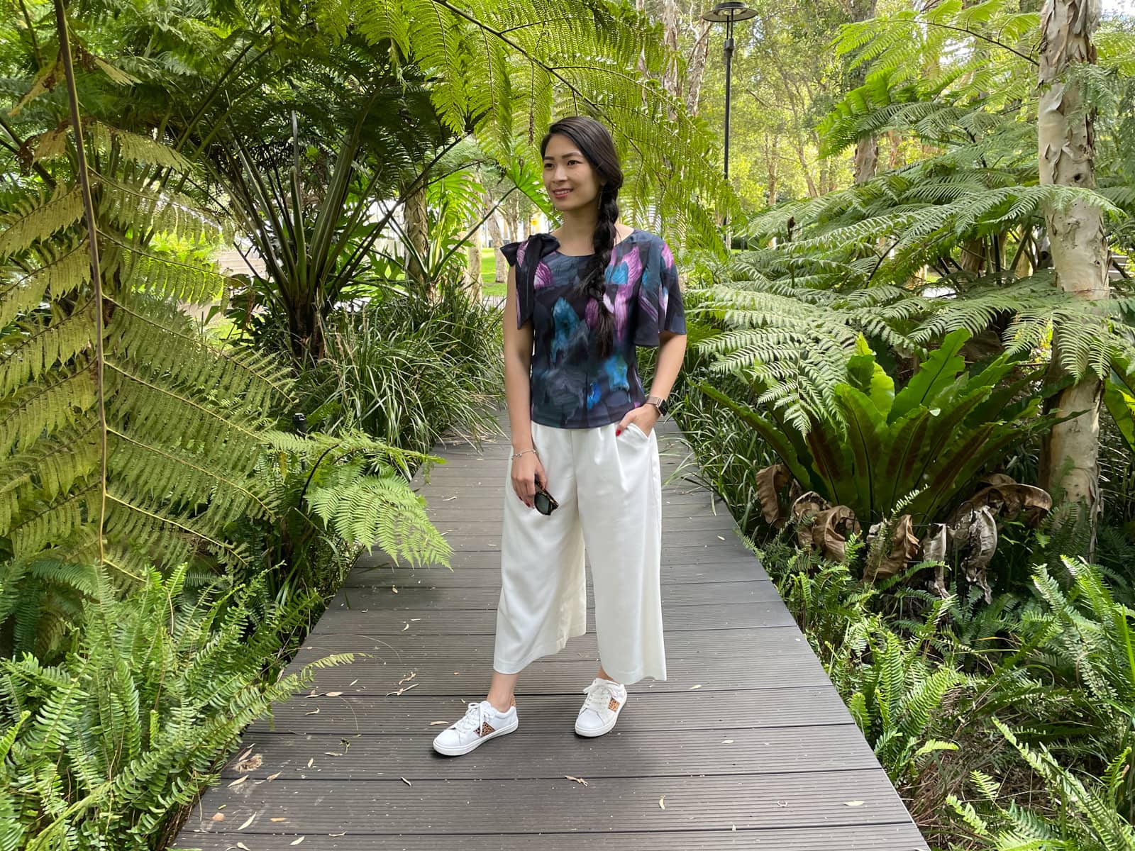 The same woman in earlier photos on this page, in the same outfit and setting. She is standing on a wooden boardwalk and has a hand in the pocket of her white pants. She is holding a pair of sunglasses in her other hand.