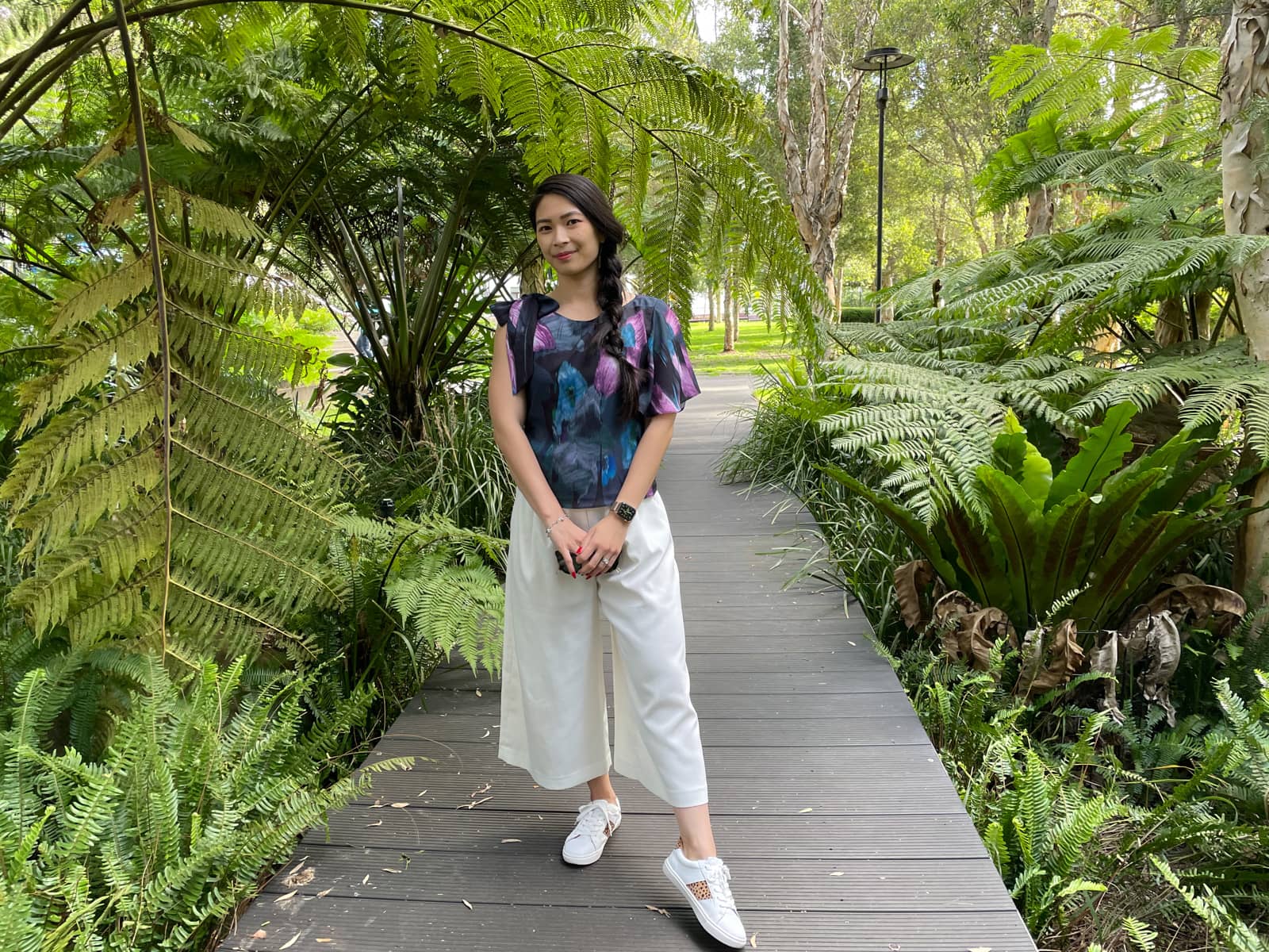 A woman with fair skin and long dark hair braided loosely in a side braid. She is wearing a patterned top and white culottes, and standing on a boardwalk surrounded by fern-like trees.