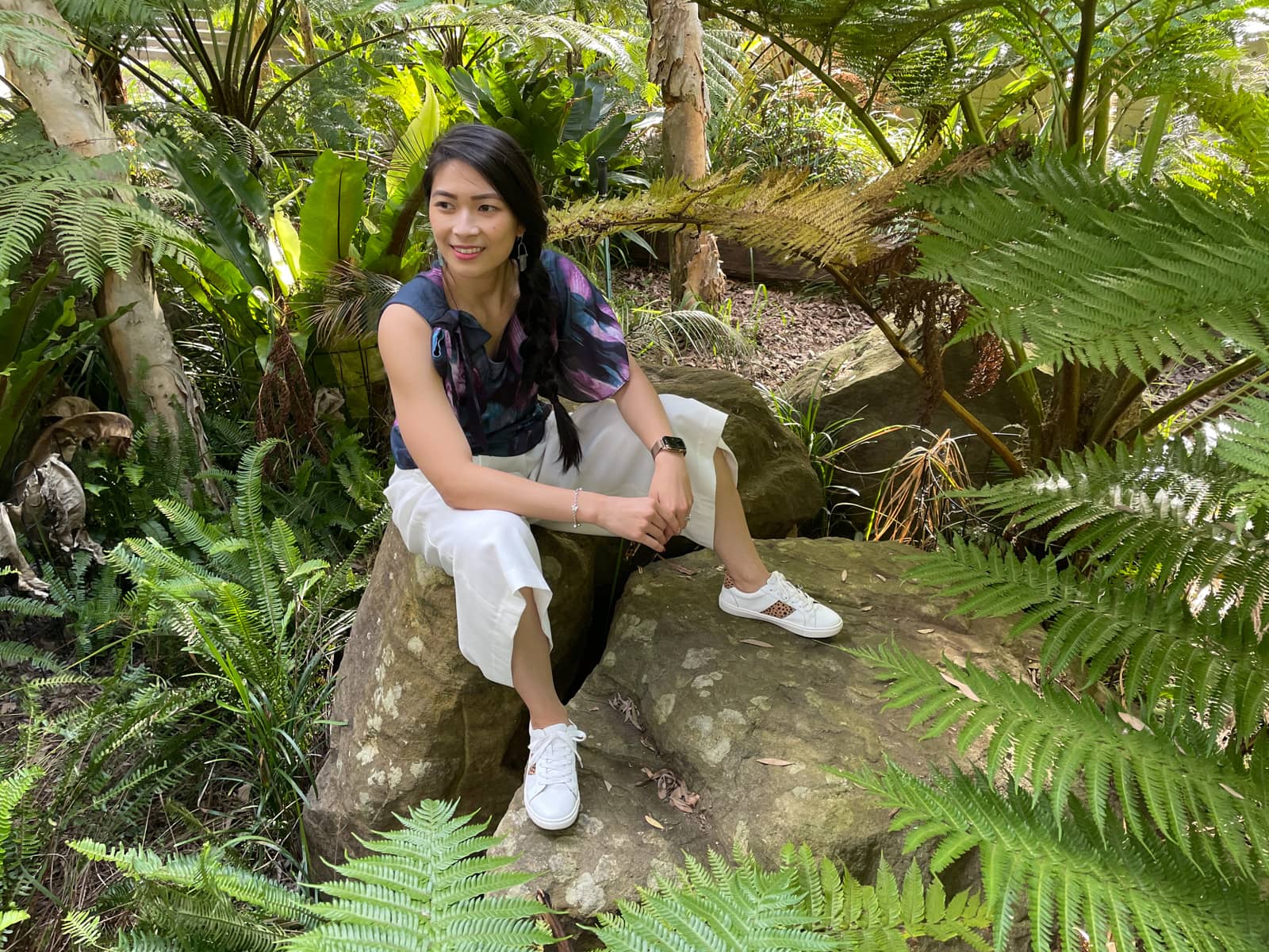 A woman with fair skin and long dark hair braided loosely in a side braid. She is wearing a dark top with a blue and purple floral pattern, white loose short pants, and white sneakers. She is sitting on a rock amongst some trees with fern-like fronds.