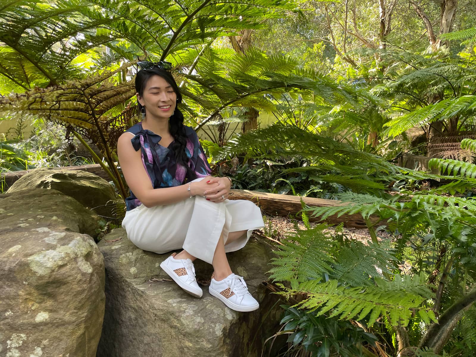 A woman with fair skin and long dark hair braided loosely in a side braid. She is wearing a dark patterned top and sitting on a rock, her eyes slightly closed