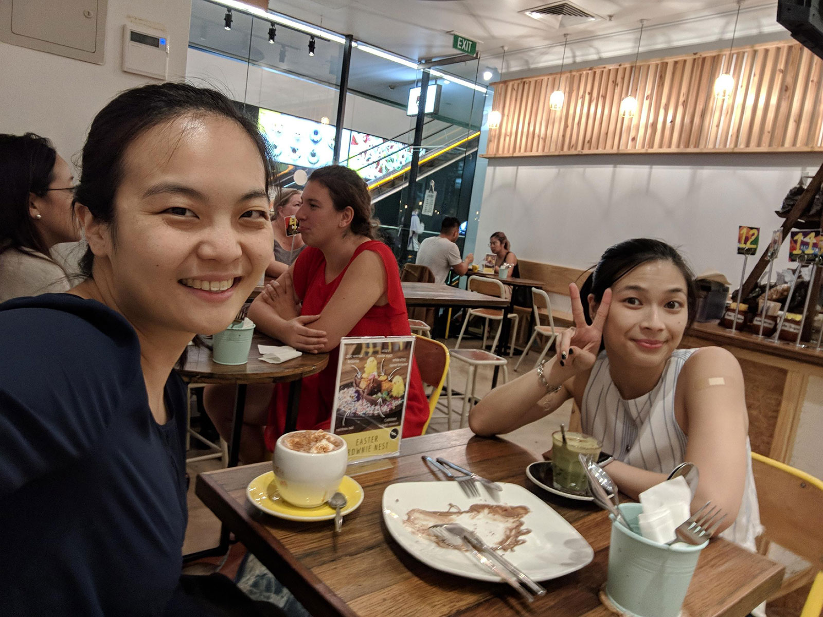 Two girls facing the camera, smiling. They are sitting at a table with two cups of hot drinks. The one further away from the camera is making a peace/victory sign with two fingers. In the middle of the table is a white plate with some chocolate sauce on it, and two sets of cutlery. They are in a cafe setting.