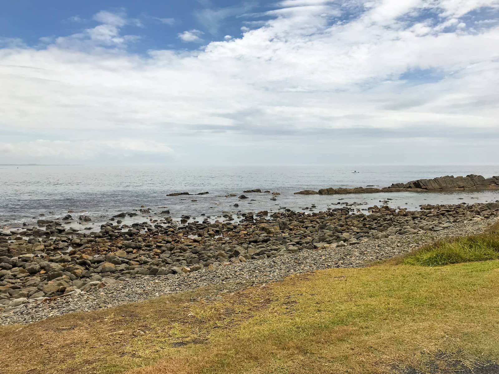 A view of rocks lining the edge of an area between grassy land and sea. The sky is blue but there are many clouds in the sky.