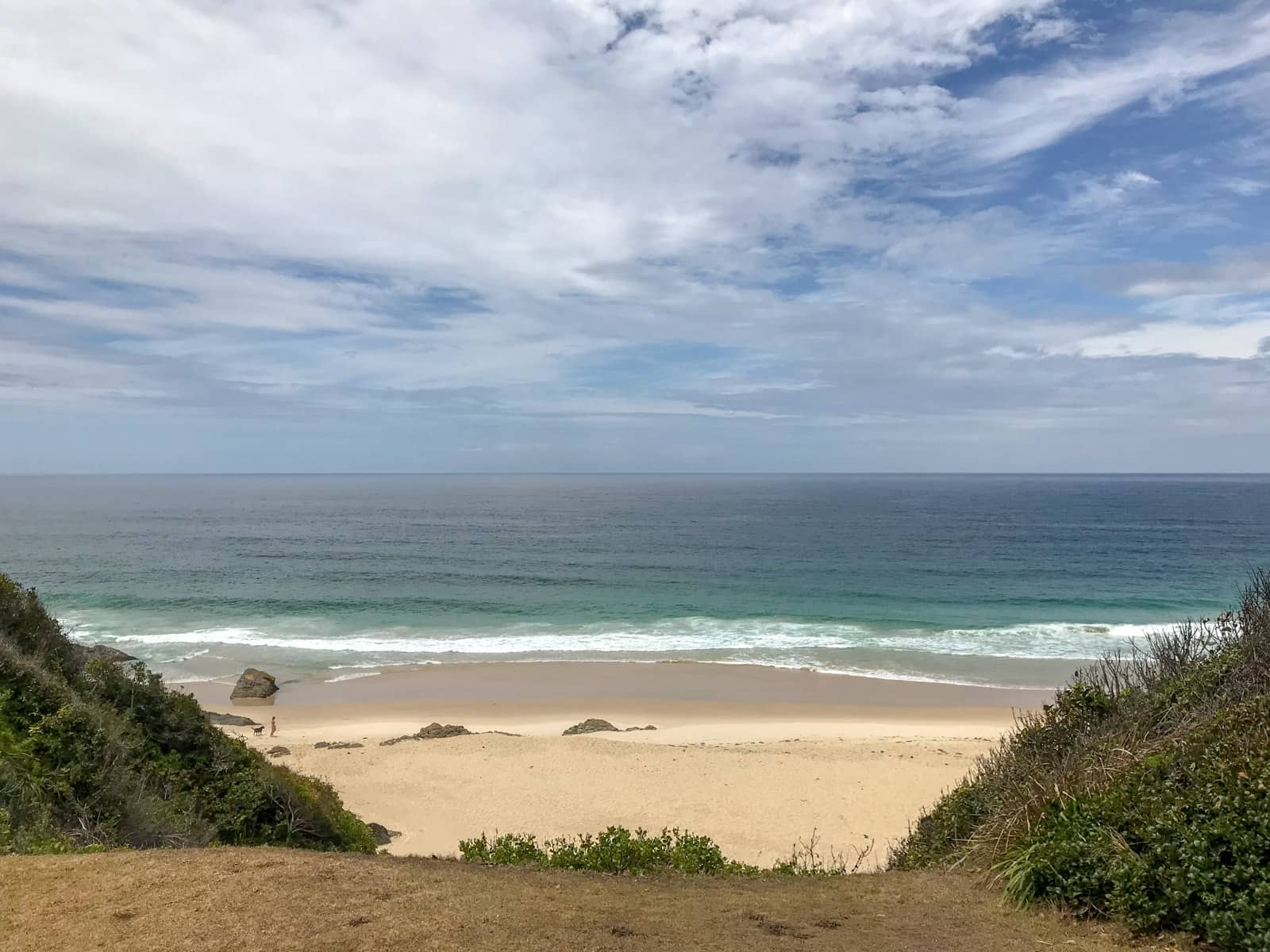 A view of a sandy beach taken from a non-sandy area further from the beach. The sky shows many clouds. In the foreground is some weedy terrain.