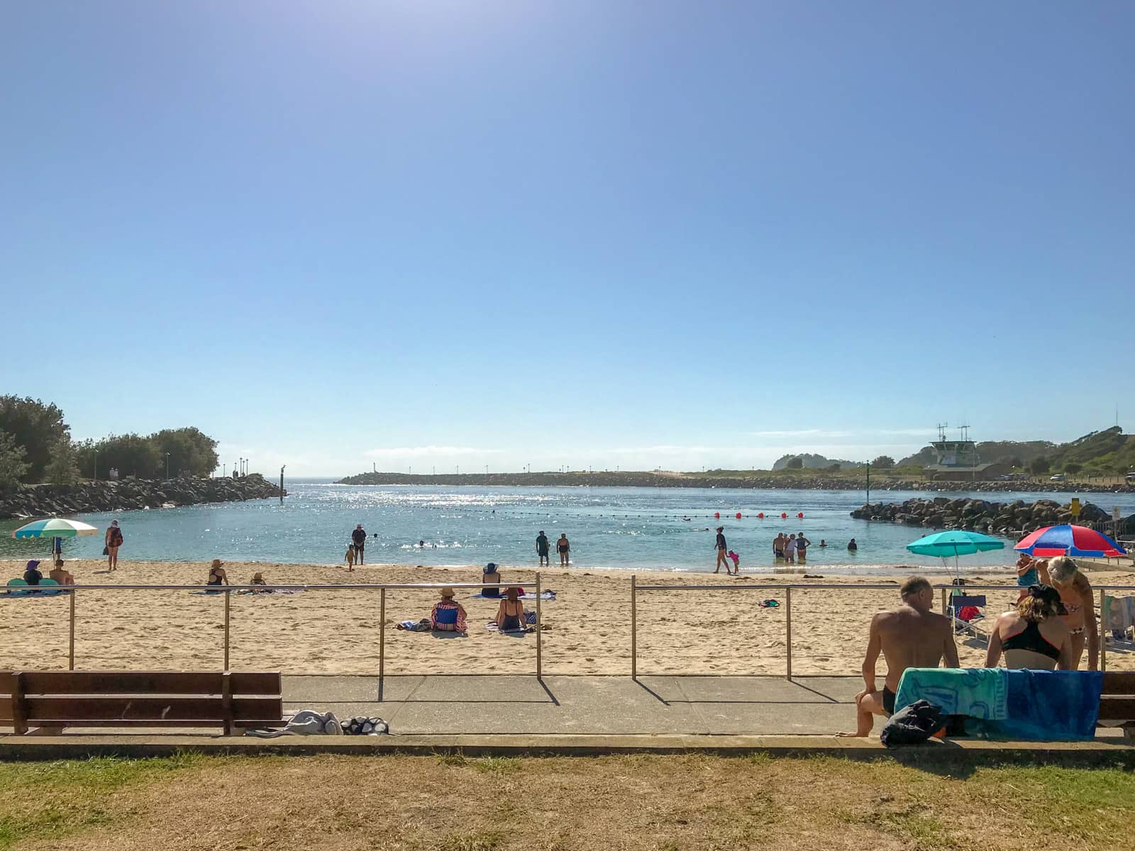 A yellow, sandy beach with quite a few people standing at the water’s edge or sitting on the sand. In the foreground is a paved path with benches at its edge, and metal railings at the edge of the path before the sand.