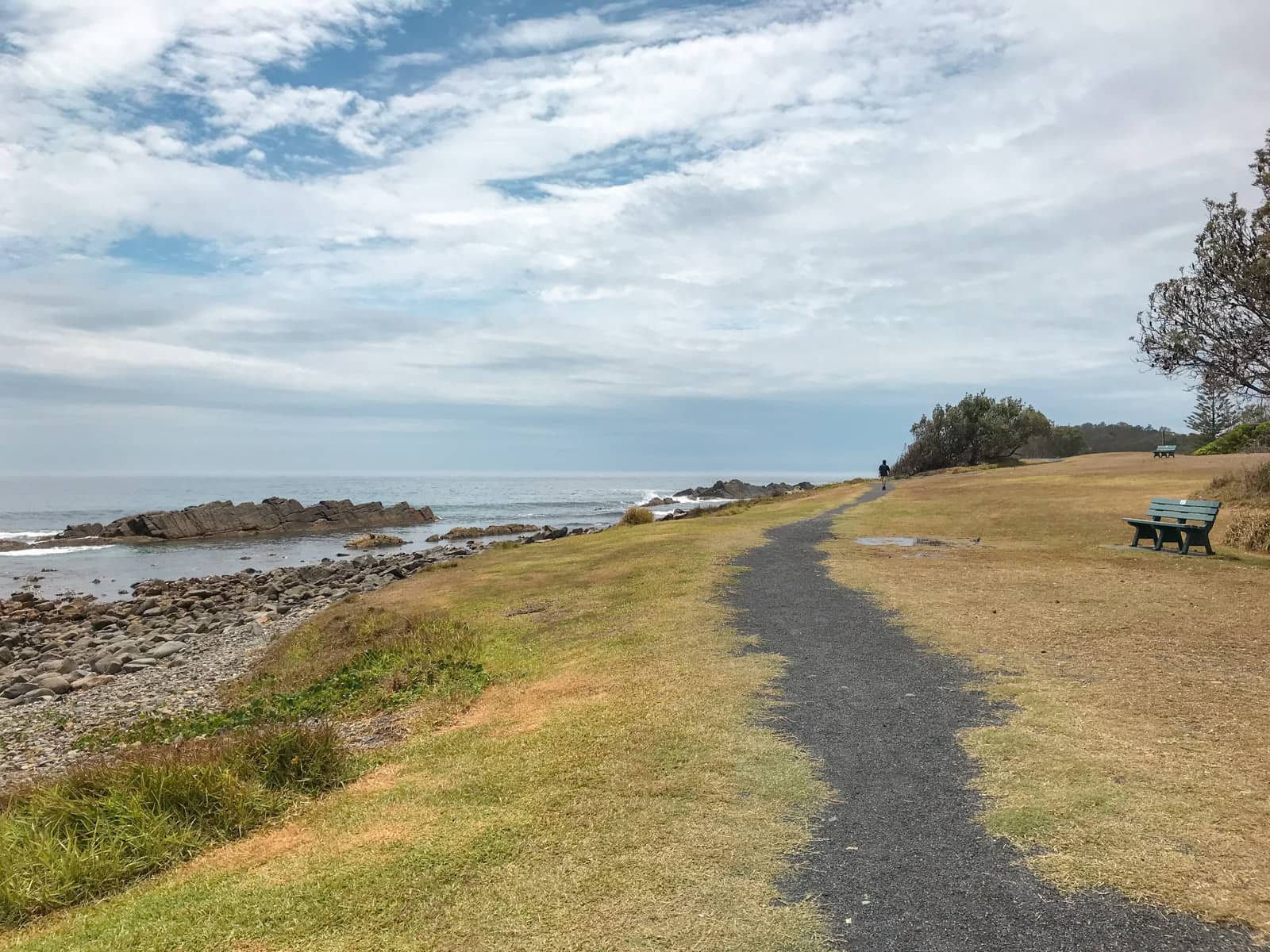 A grassy area with a grey gravel path leading into the distance. The path has ragged edges. On the left is a rocky beach leading to the sea, and on the right is a wooden bench in the foreground. A man can be seen walking on the path in the distance