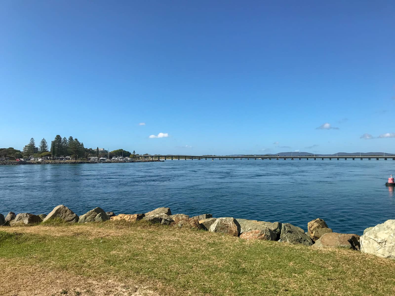 A very blue body of water with a road bridge in the background, appearing horizontally. In the foreground is grass and large rocks lining the edge of the grass before the water.