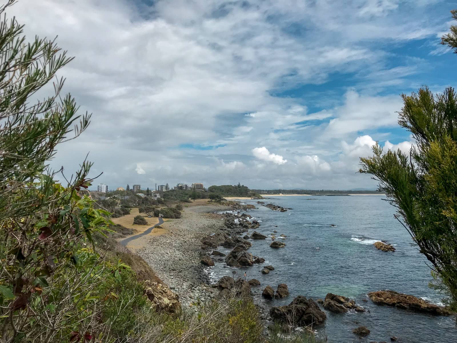 A view between some trees, at a high elevation. Some buildings can be seen in the distance, but in the foreground the path from the previous photo can be seen, with a greater view of the sea on the right.