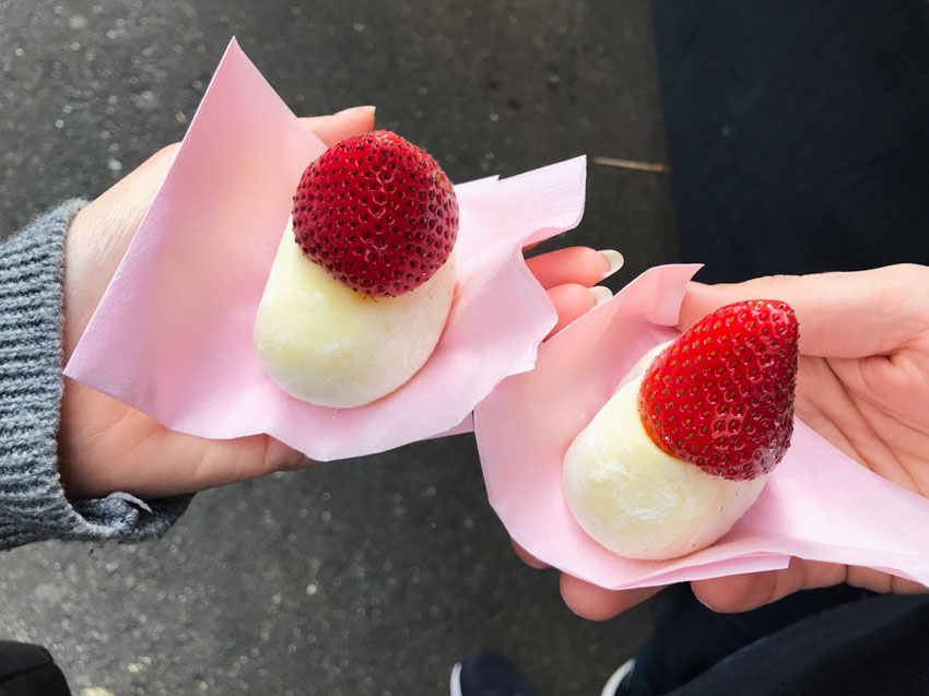 Two separate peopleâ€™s hands holding pale yellow mochi with a single strawberry on top of each, placed upside-down. The mochi sits on pink napkins.