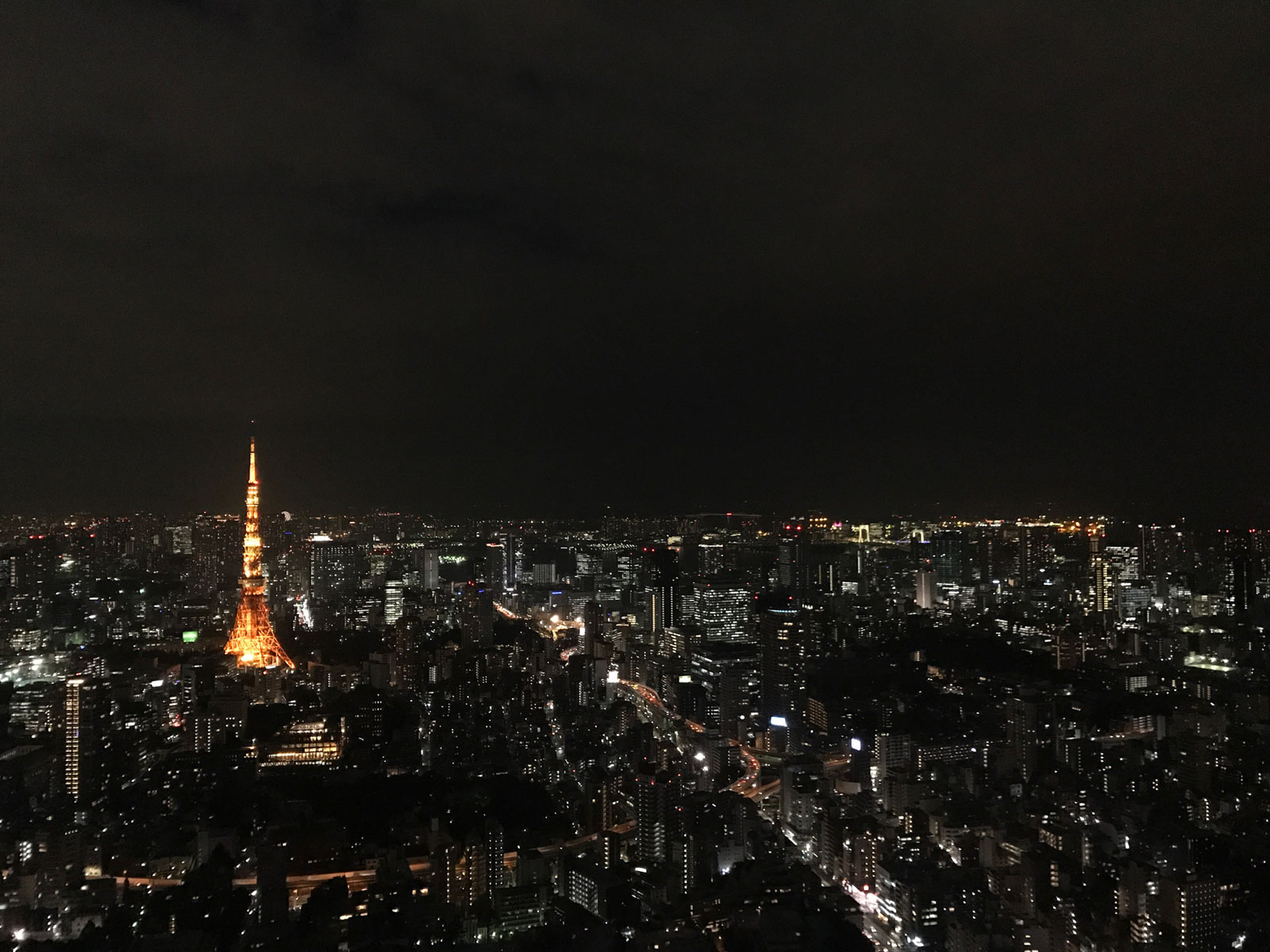 A city at night, with many buildings with lights on. One brightly lit tower with yellow lighting stands out amongst the other buildings