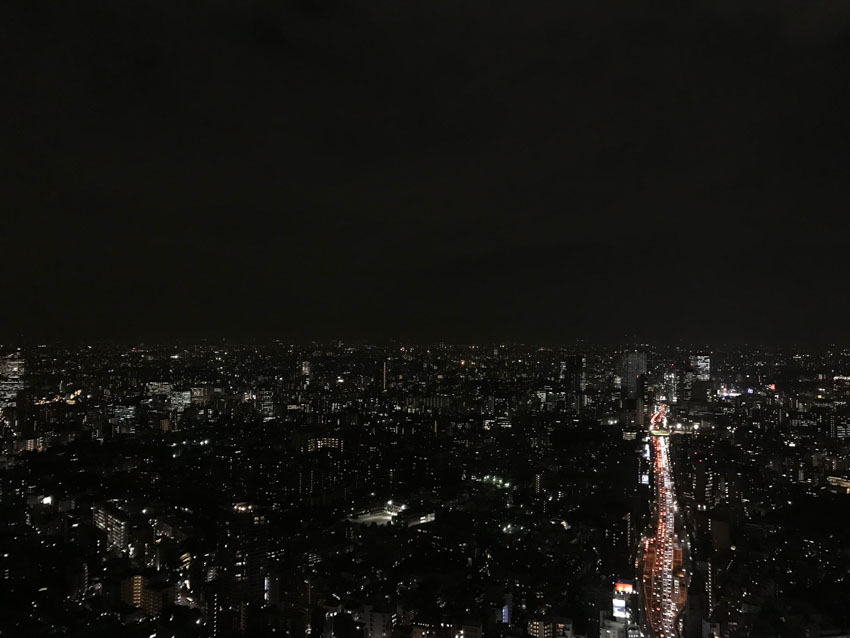 A wide view of a city at night from a high altitude. A long line of different coloured lights shows a road of traffic one side of the photo, moving into the distance.