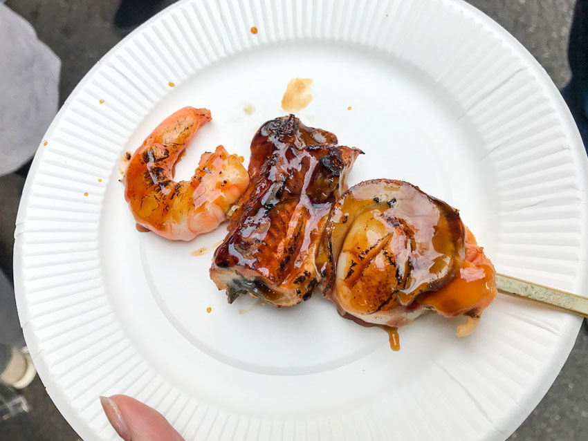 A white paper plate with a skewer, the skewer has a prawn, piece of eel, and scallop (from top to bottom), and is covered in dark brown sauce