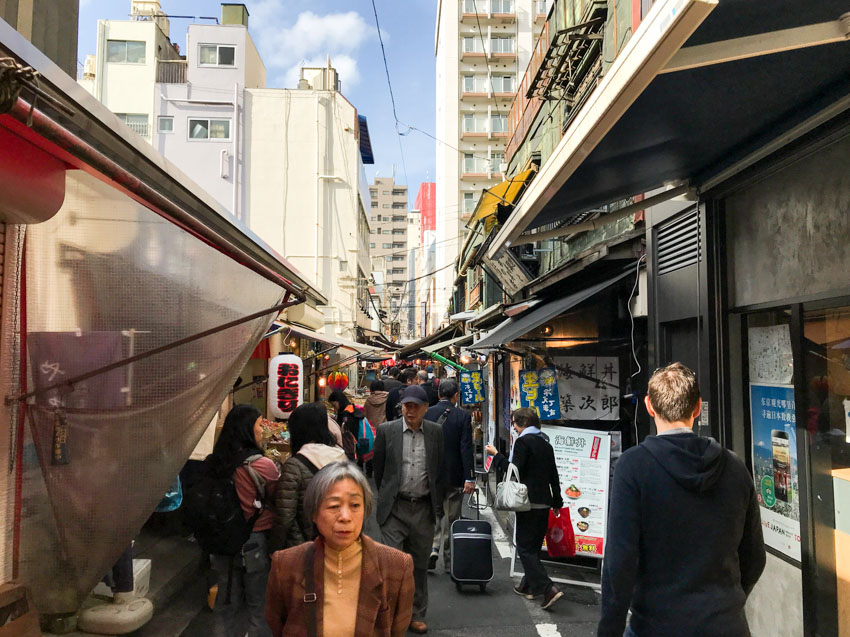 A market during the day. Some Japanese signage can be seen on the right side. Many people are walking through the market ahead.