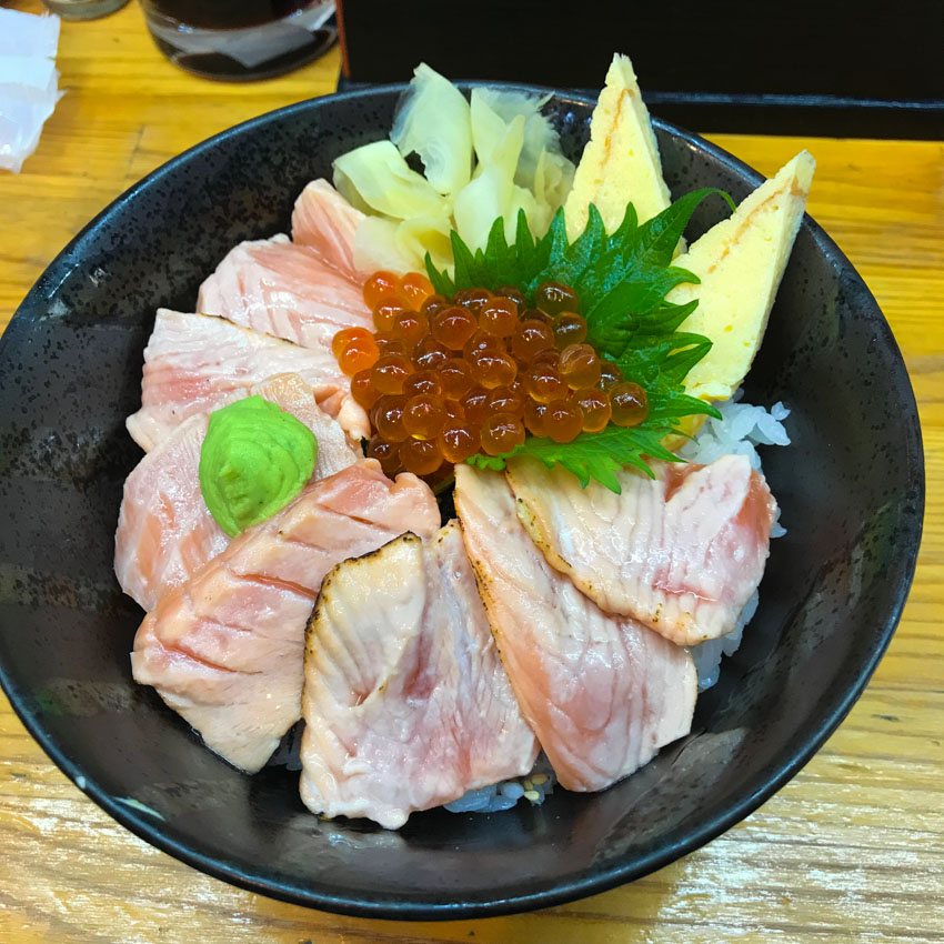 A black bowl containing rice with lightly grilled salmon. Itâ€™s topped with a pea-sized piece of wasabi, orange cod roe that looks like translucent little globes, and yellow pickled ginger.