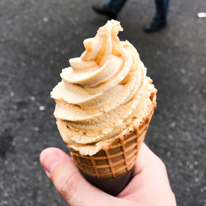 A hand holding a waffle ice cream cone, filled with a swirl of light brown ice cream with golden brown caramel sauce through it