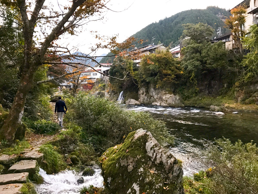 The river with some houses visible, and Nick facing away from the camera, walking along the path