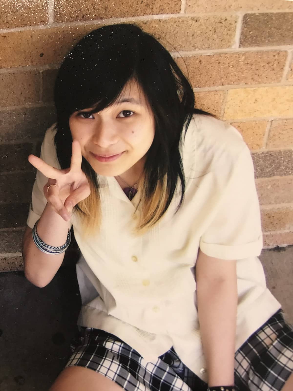 A girl in a school uniform holding her two fingers up in a peace sign. She has short dark hair with blonde ends