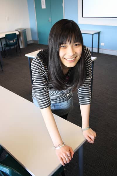 A young woman leaning forward on a desk while standing. She’s smiling and her hair is dark with a straight fringe. She is wearing a dark grey and black striped top.