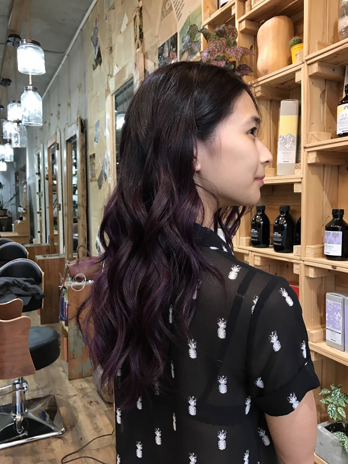 A woman with a black shirt, looking away from the camera. She is standing in a hair salon and her hair is freshly waved with purple colour throughout.