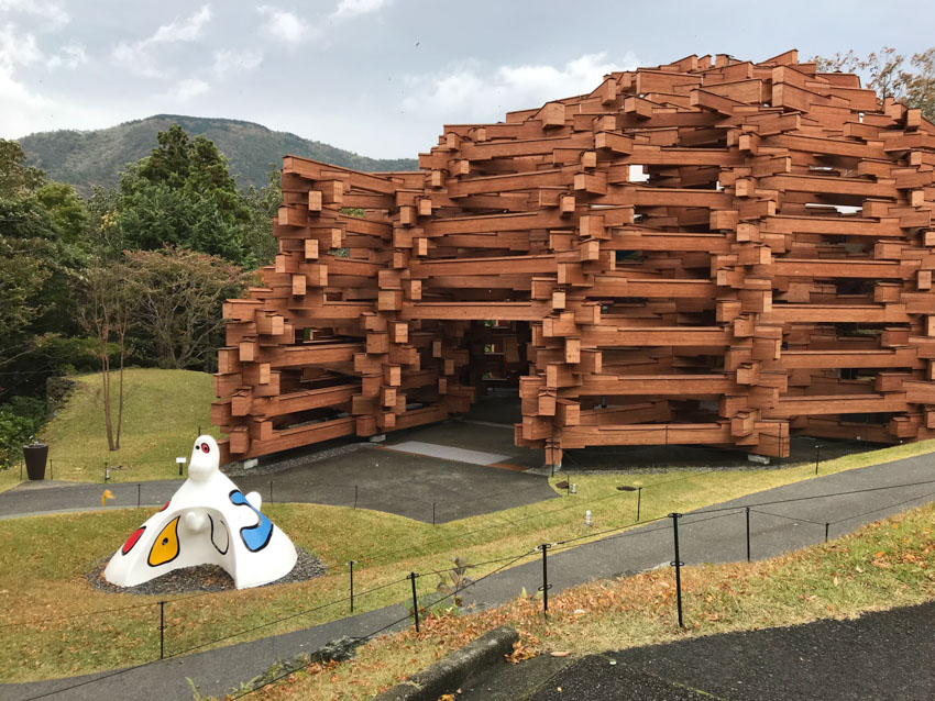 A dome structure made from giant wooden slats, accompanied by a small white cone-shaped white sculpture with a small head and painted eyes with red, yellow, and blue patches