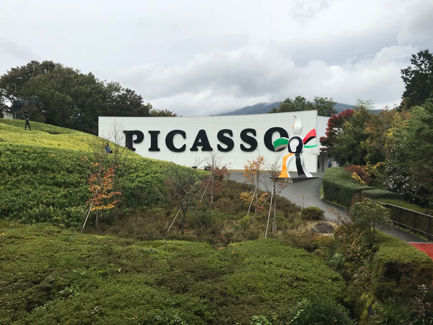 A large curved white building resembling a wall, with “Picasso” printed on it in uppercase, amongst the green bushes