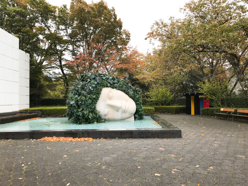 A large sculpture of a head on its side with green foliage as hair, sitting in a man-made shallow pool