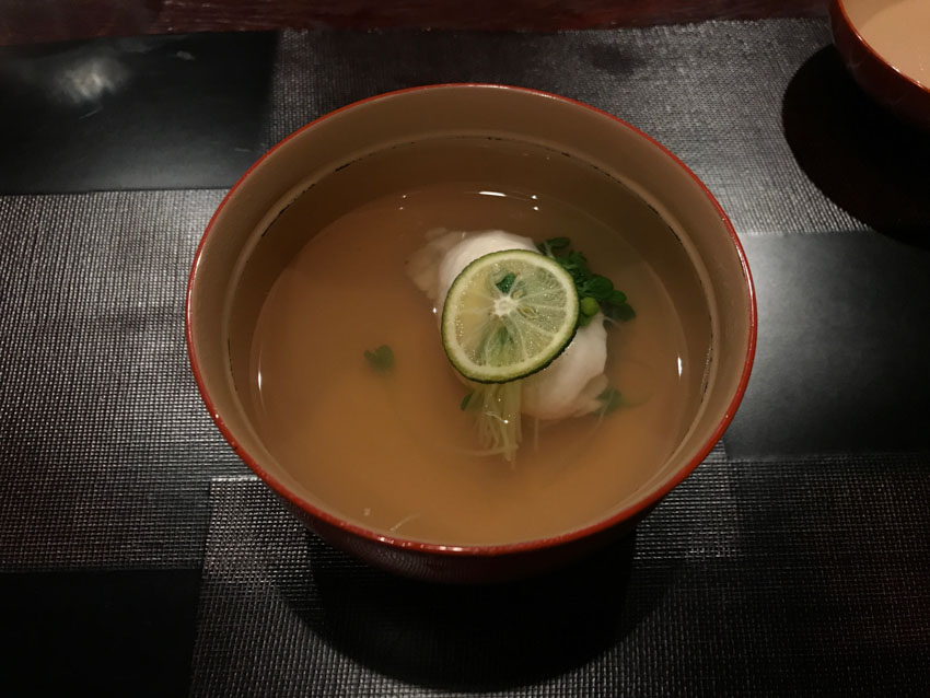 Top-down view of a bowl with boiled seasoned water and a piece of cod topped with radish sprouts and a lime