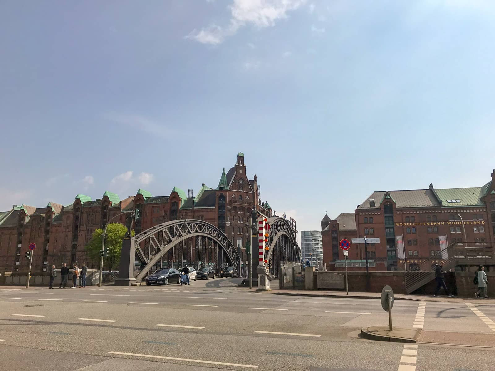 A small bridge with arches on either side, as seen from across the road that is perpendicular to it. The sky shows it’s in the afternoon.