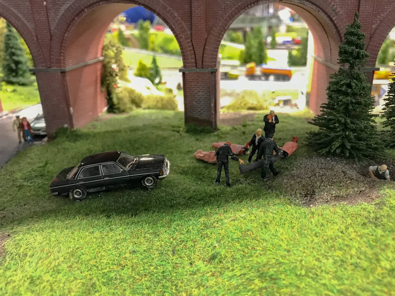 Minature figurines dressed in black, near a black car, under a bridge. They are dragging what appears to be figurines wrapped in paper bag.