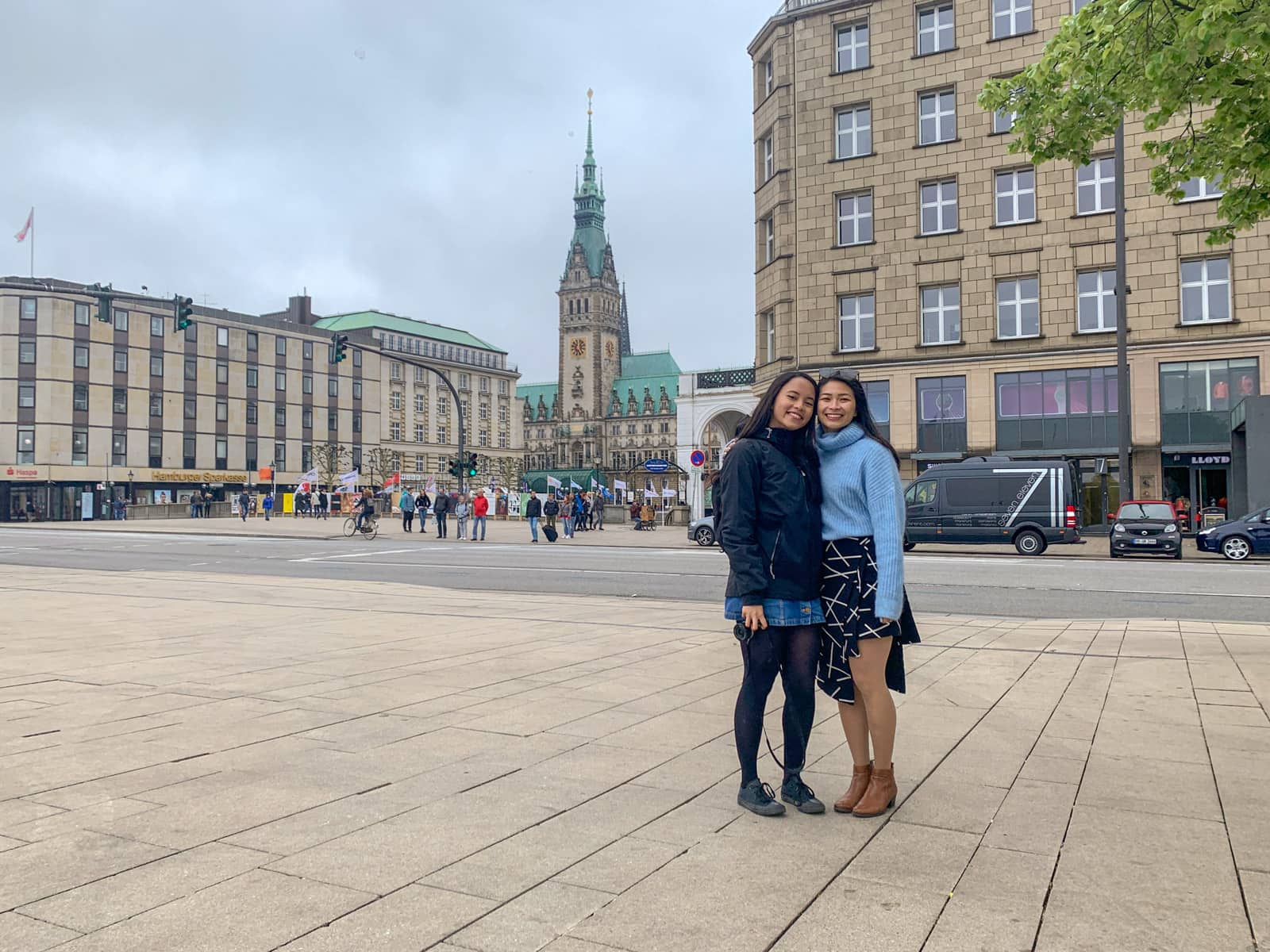 Two women smiling, with their arms around each other. Both have dark hair. One is wearing a striped coloured top and a black jacket with a blue skirt, the other is wearing a light blue sweater with a black-and-white patterned skirt. They are in a street setting with a clock tower in the background.