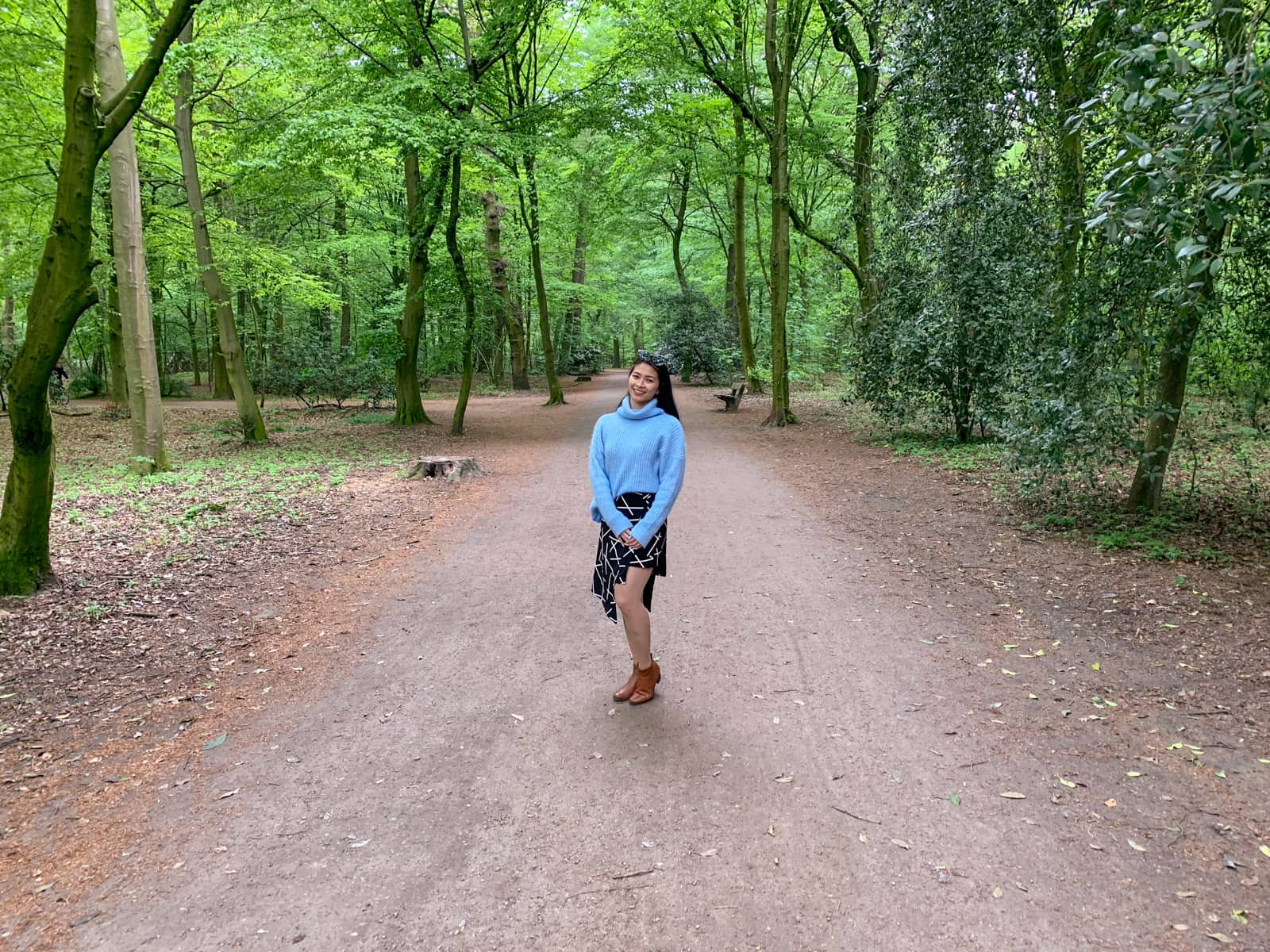 A woman in a light blue sweater and black-and-white patterned layered skirt. She’s standing on a dirt path in a forest-like area with a lot of lush green trees.