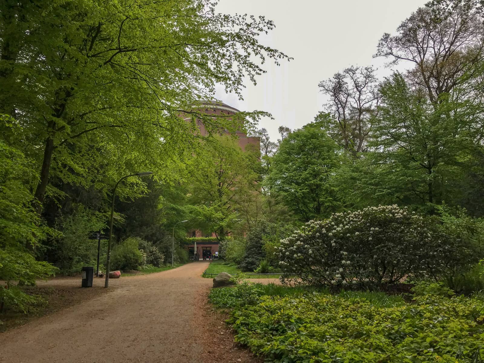 The inside of a large garden. In the background is a large brown building, partially covered by trees in front of it. The trees have many leaves but their branches are still visible. A dirt path leads in the direction of the building. To the right of frame are some planted shrubs.