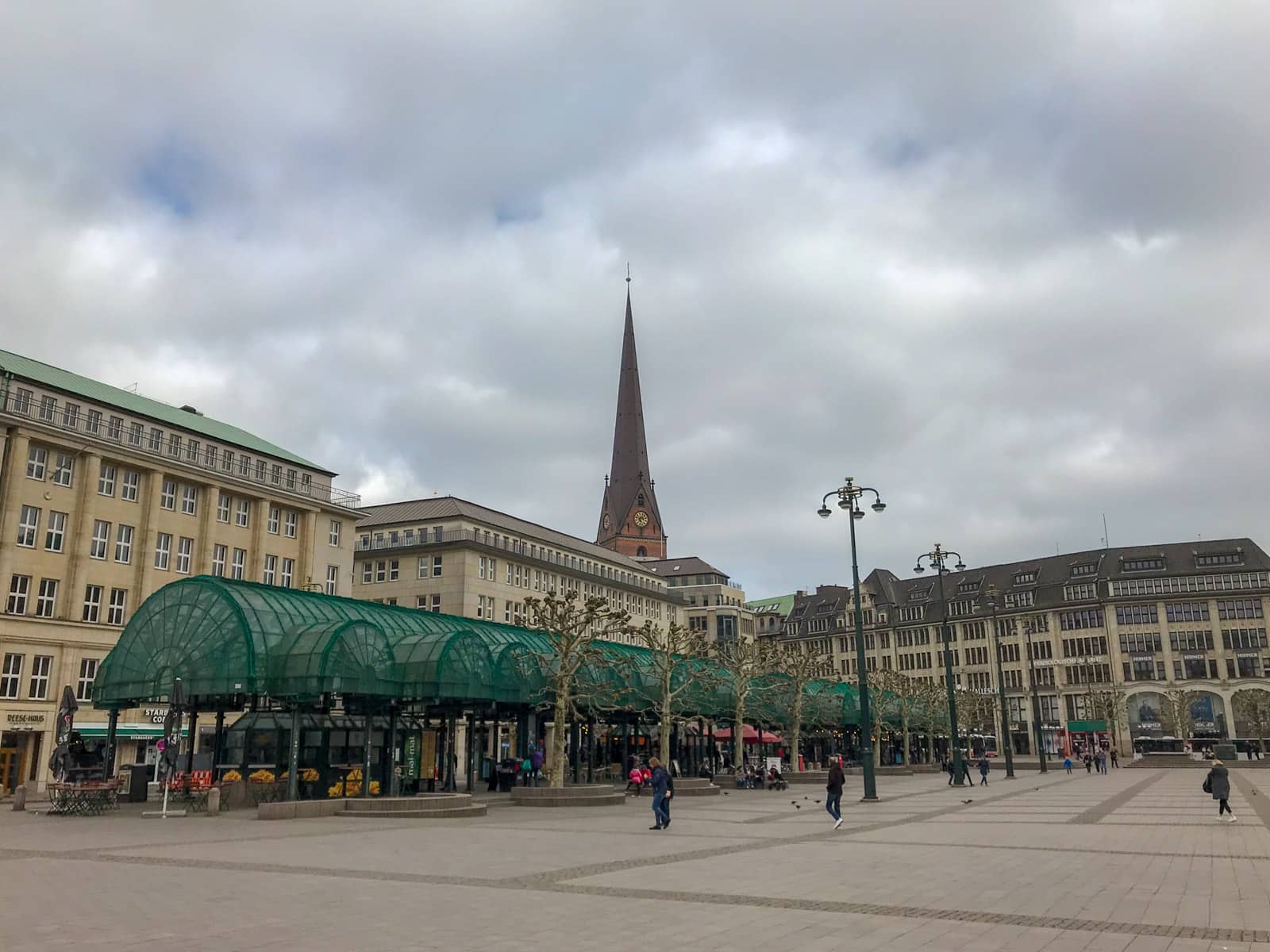 A city square/plaza, with a covered area and a tower in the background behind some buildings