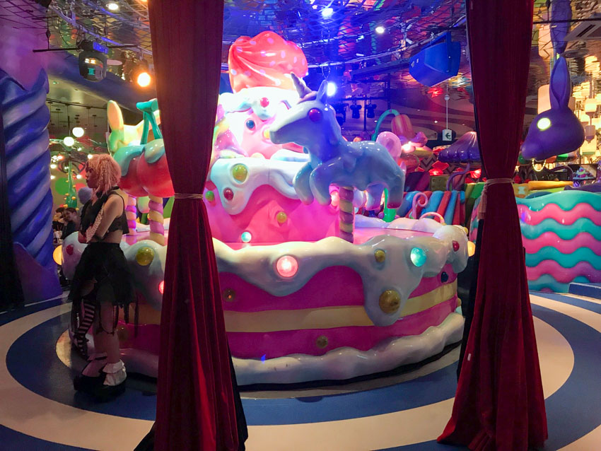 Two tied dark red drapes in the foreground. A waitress in a black frilly dress with light curly hair stands in the background, next to a giant sculpture of a cake with horses on it