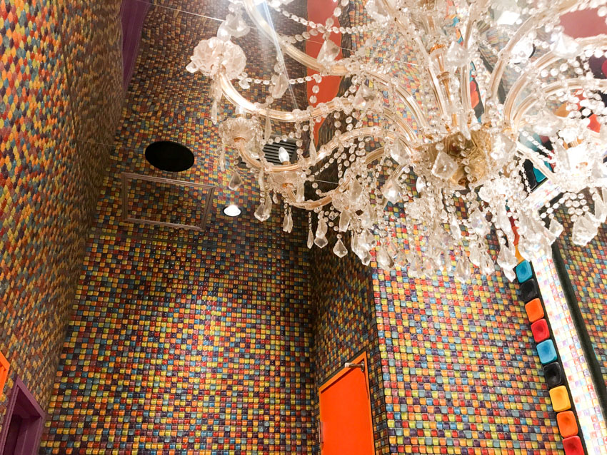 Walls in a bathroom covered with clourful square tiles, with a classic chandelier hanging from the ceiling.