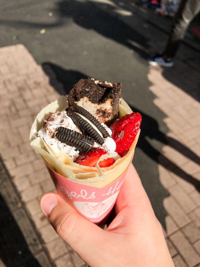 A manâ€™s hand holding a rolled-up crepe containing strawberries, Oreo biscuits and whippe dcream