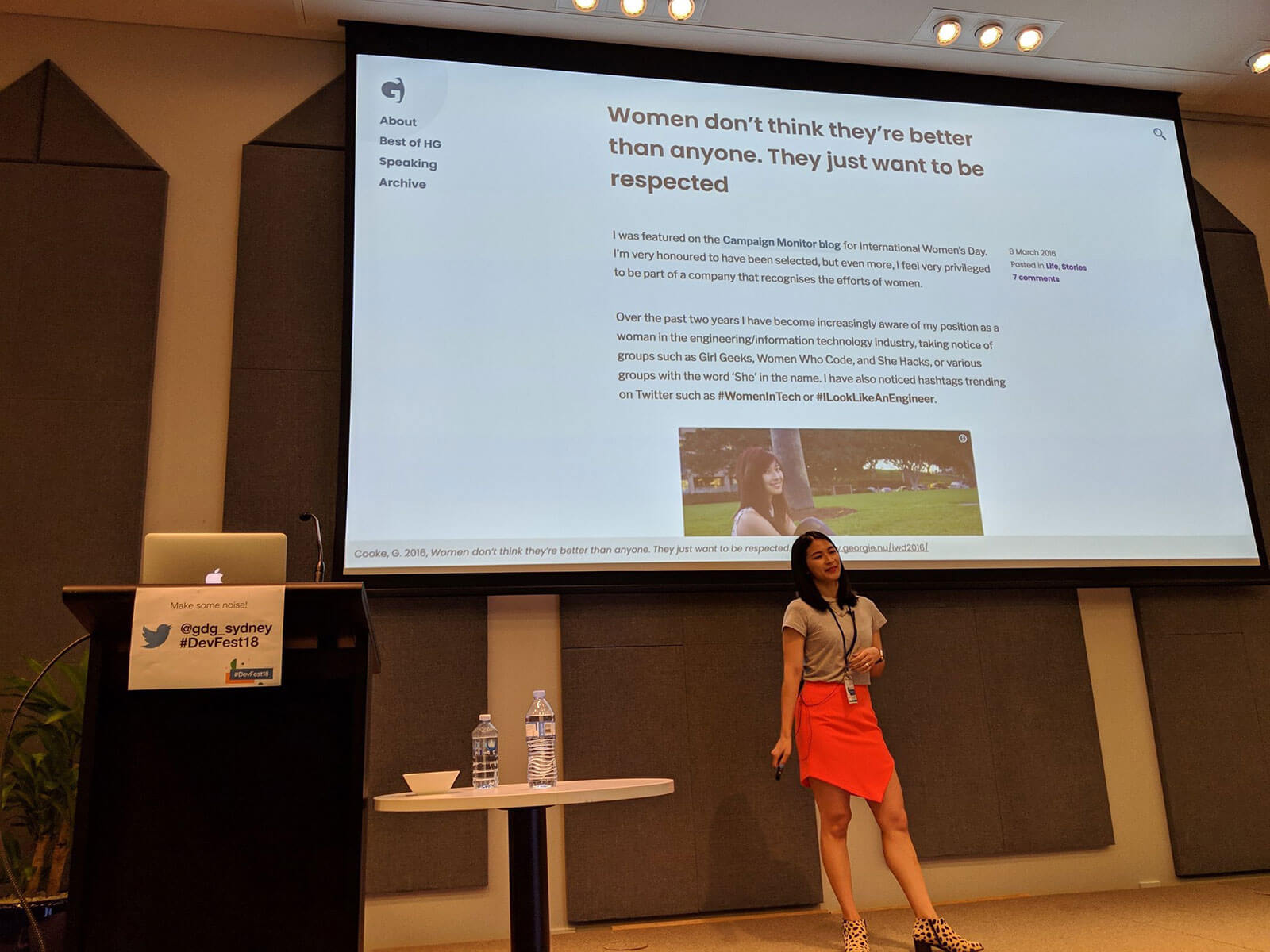 A woman in a grey t-shirt and orange skirt, speaking on stage. Behind her is a screen with a screenshot of a blog post on it