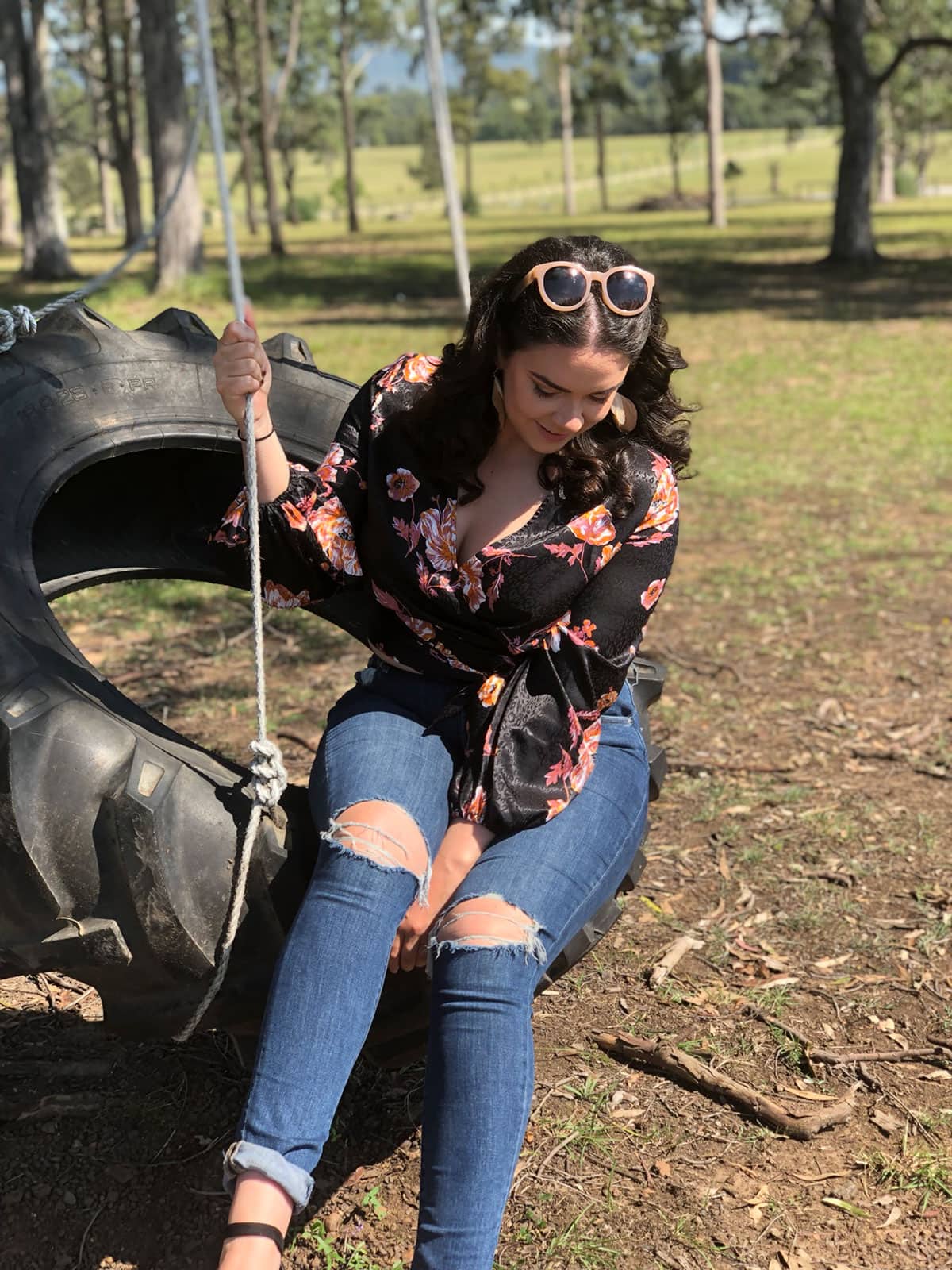 A woman with curly dark hair, wearing blue ripped jeans and a black floral blouse sitting on a tyre swing