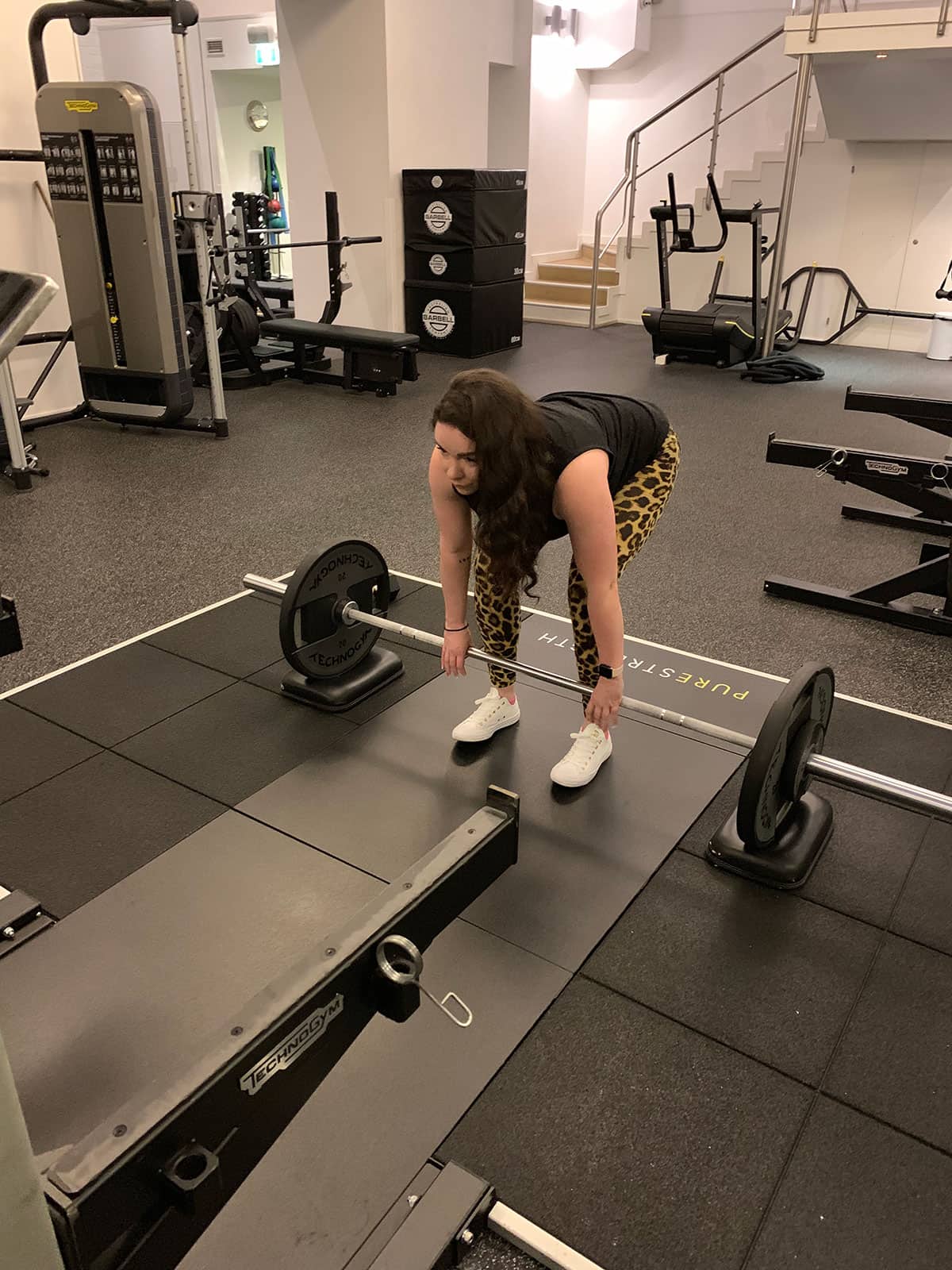 A woman with curly dark hair, dressed in activewear, preparing to do a conventional deadlift in a gym