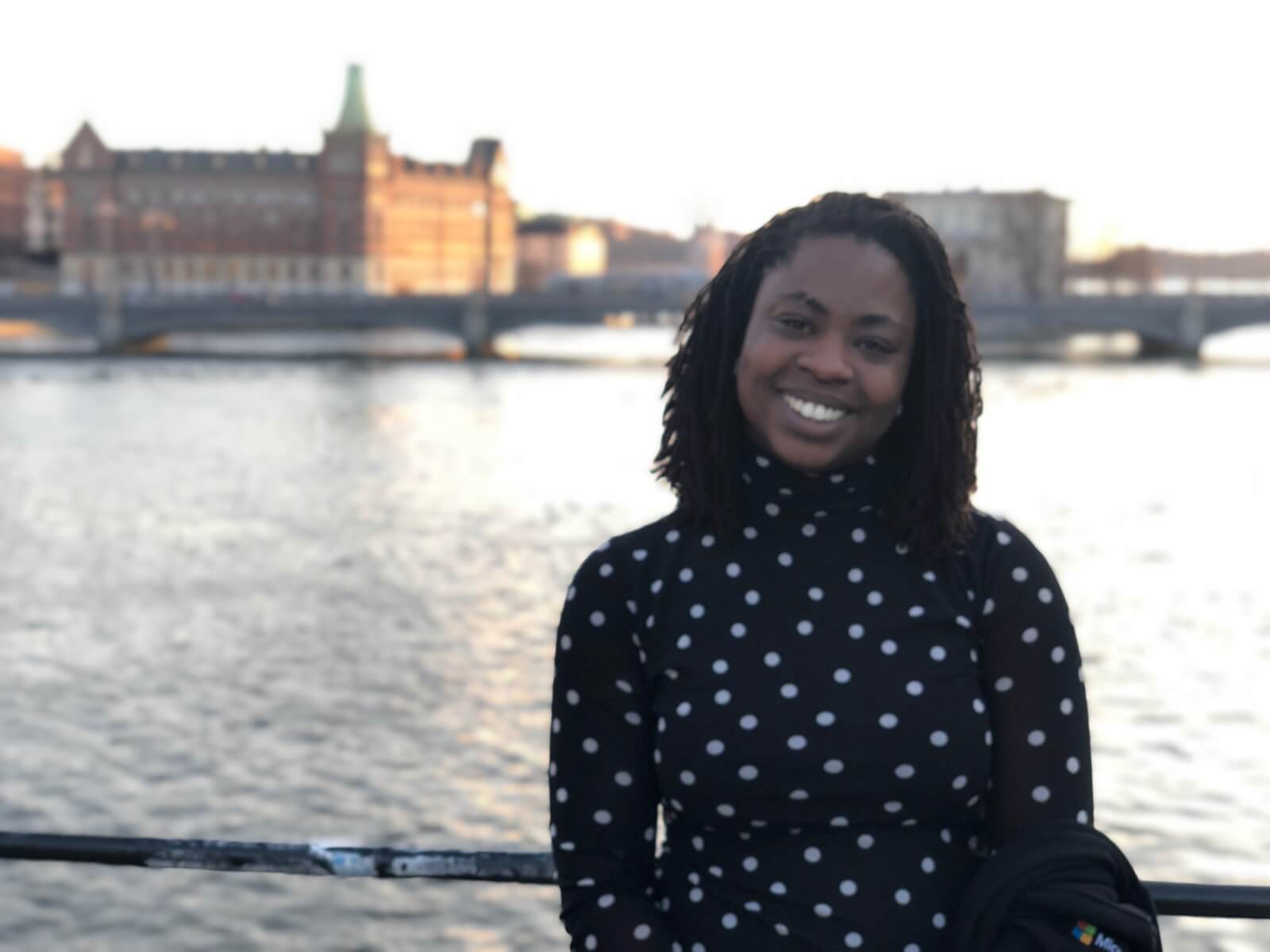 A dark-skinned woman with dark curly hair and in a black and white spotted top, smiling. There is a river in the background and a historic building in the distance