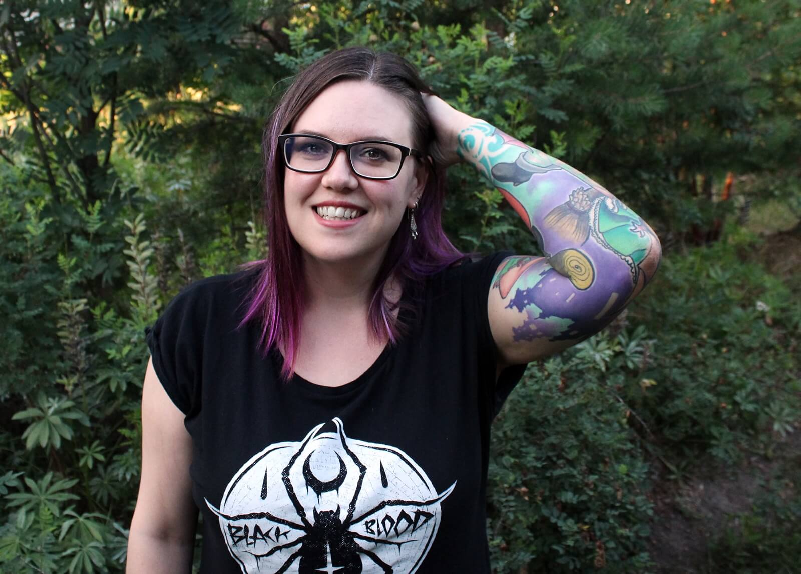 A light-skinned woman wearing glasses and a black t-shirt. She has purple and brown hair. Her hand is held to her head with her elbow out to the side. Her arm has many tattoos.
