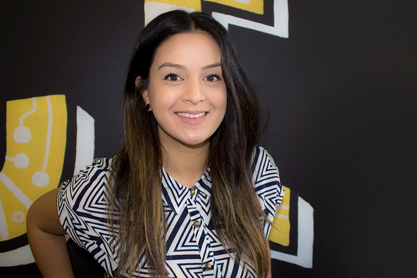 A girl smiling, wearing a triangular patterned black-and-white collared top. The backdrop is mostly black with some yellow and white shapes resembling big letters that are cropped out of frame.