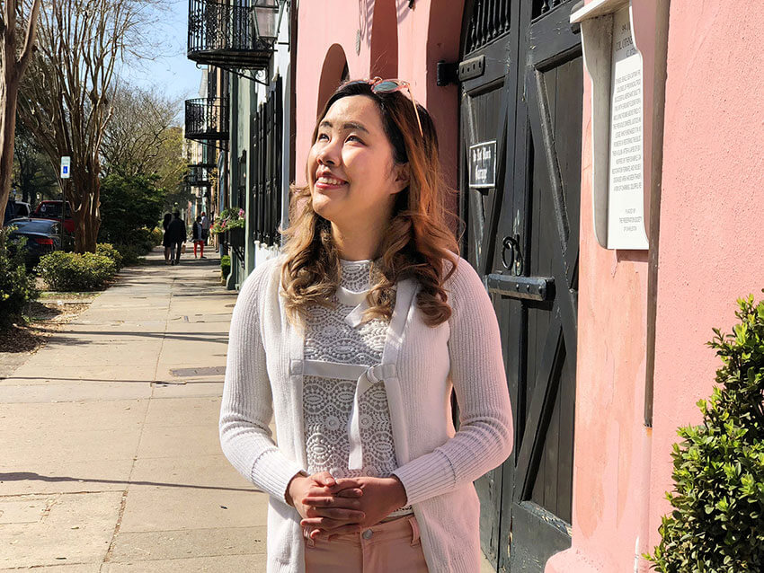 A girl with medium-length wavy brown hair with blonde highlights. She is wearing pink pants and a white cardigan and top, standing on a sidewalk by a pink building. She is looking upwards and smiling.