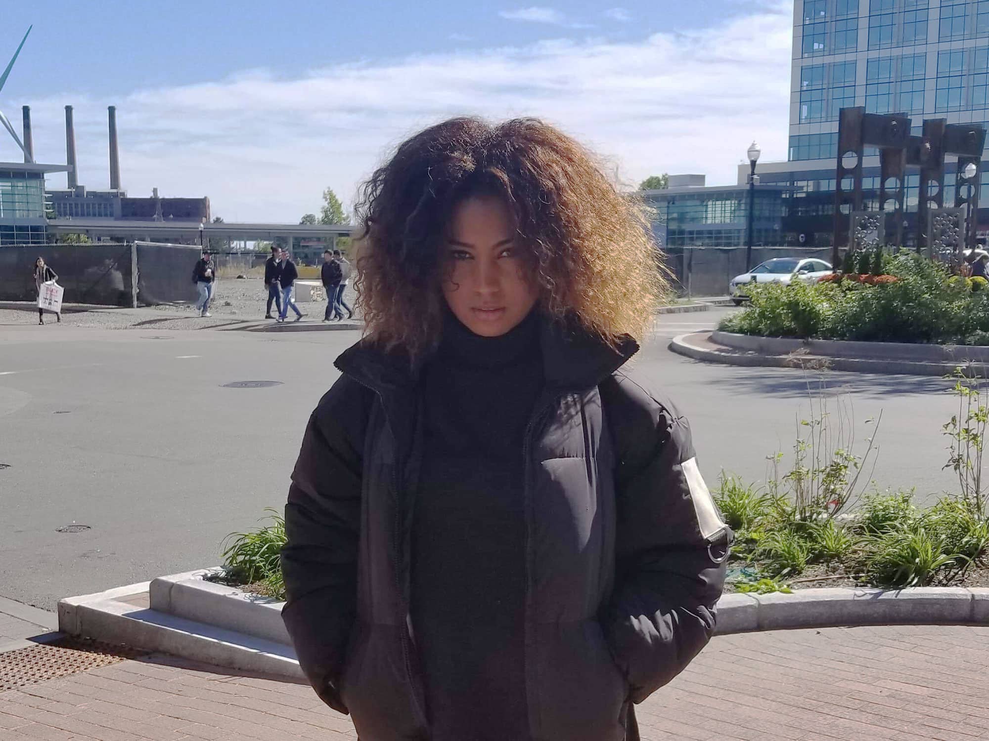 A woman with dark skin and curly dark hair with highlights, wearing a black puffer jacket. She is looking fierce and standing in an industrial area that resembles a parking lot, with a high-rise building in the background