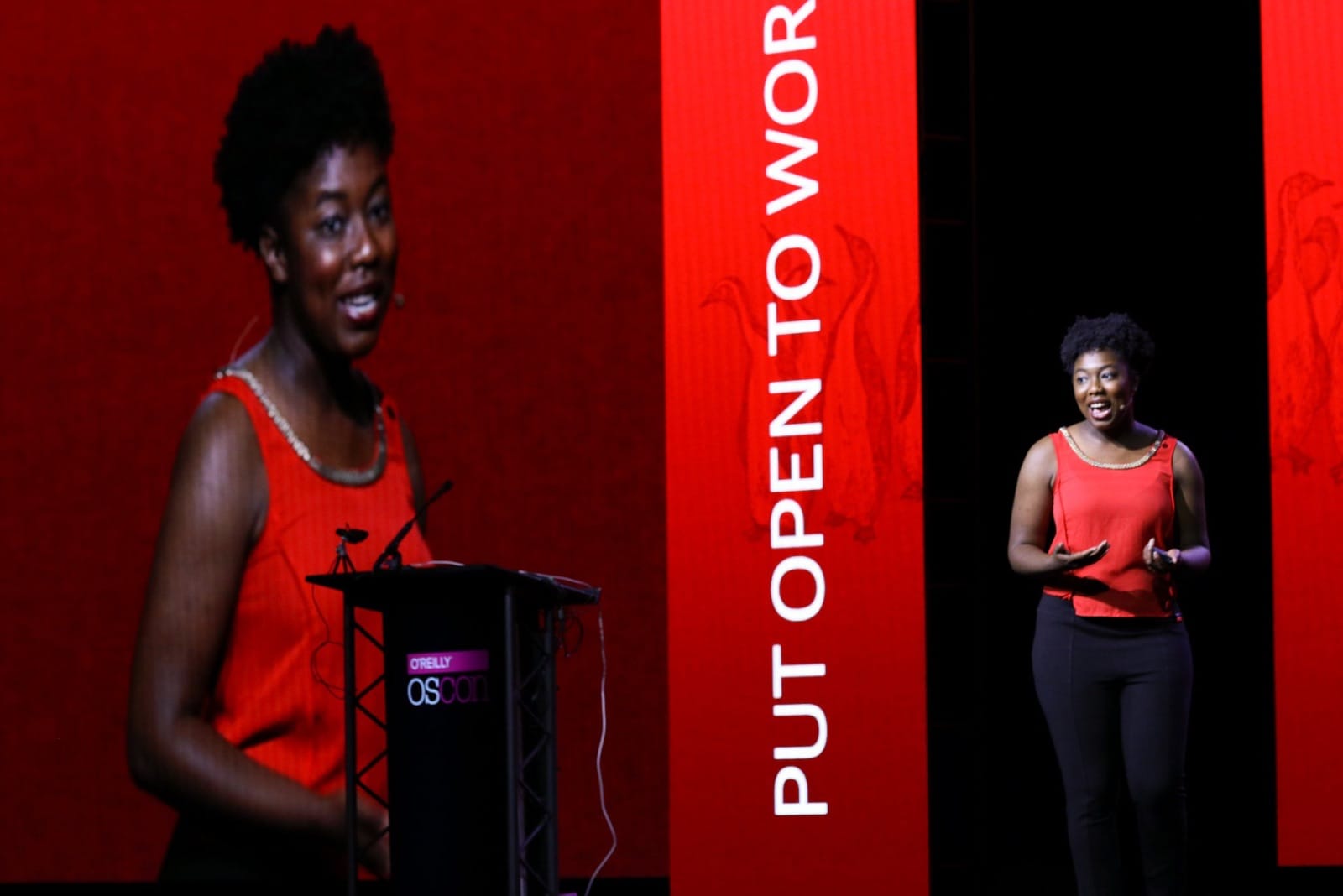 A woman with dark hair and dark skin, wearing a red top, standing on a stage near a podium